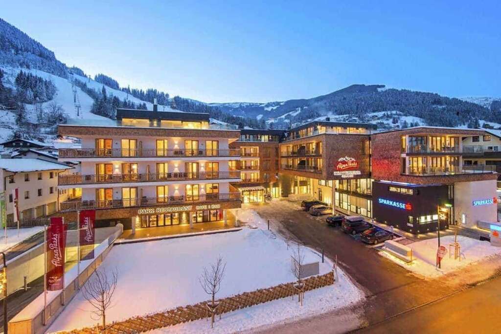 Beleuchtete Appartementhäuser an einer verschneiten Straße in der Dämmerung, dahinter Skipiste