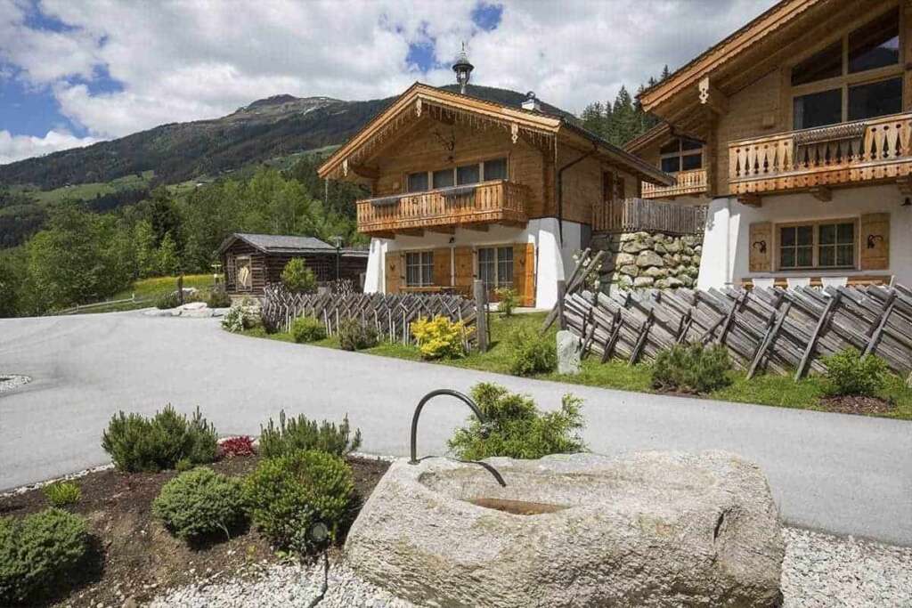 Holzchalets mit Balkonen an einer Straße, vorne ein Steinbrunnen, dahinter bewaldete Berge