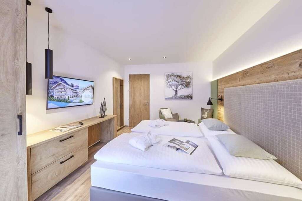 Helles Doppelzimmer mit Altholzwand hinter dem Bett, gefalteten Bademänteln und Wandfernseher
