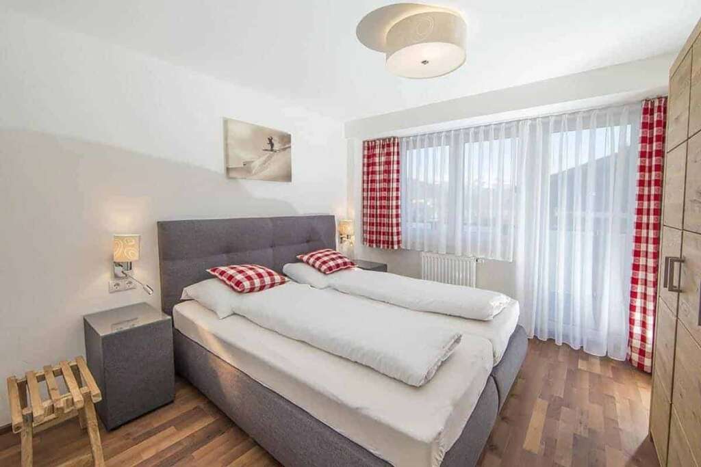 Schlafzimmer mit grauem Boxspringbett, rot-weiß karierten Kissen und Vorhängen und großem Fenster