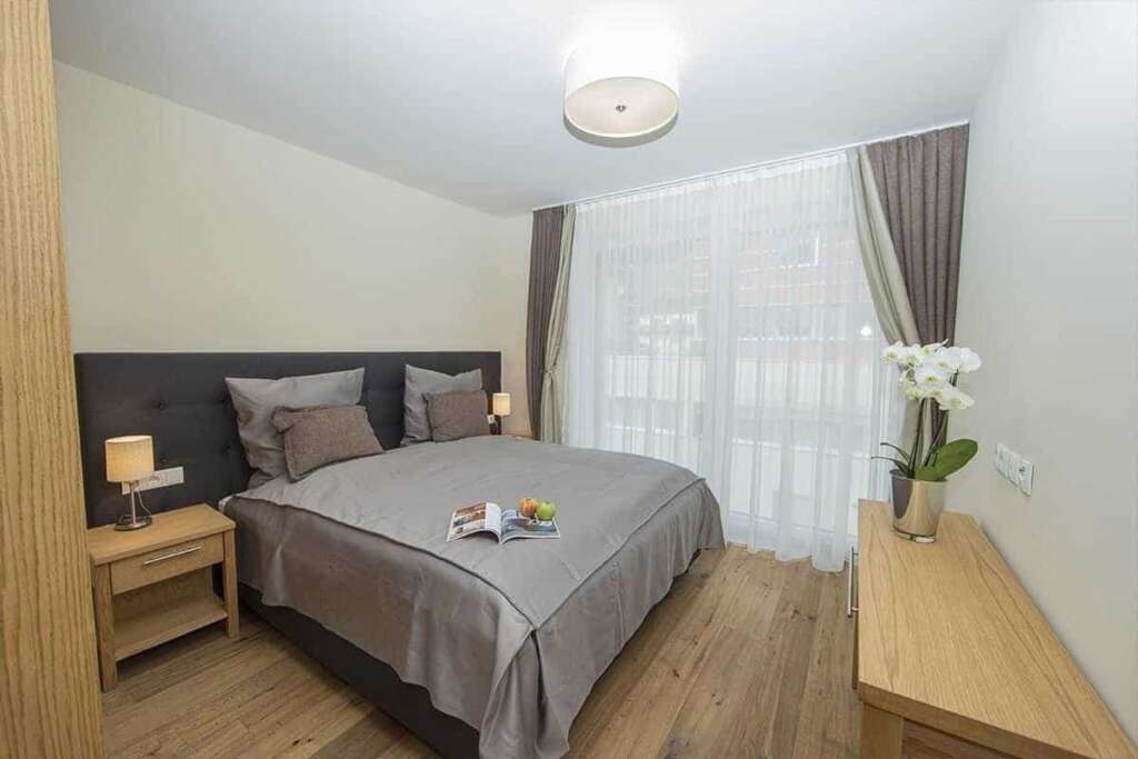 Helles Schlafzimmer mit grauem Doppelbett, Holznachttisch, Orchidee und bodentiefem Fenster