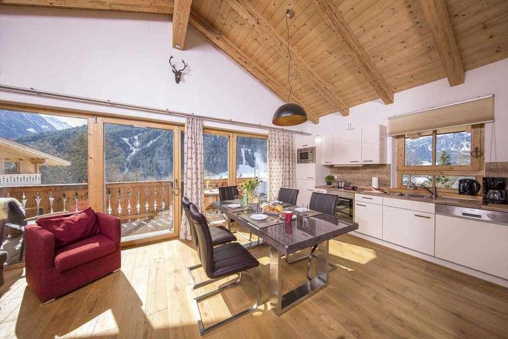 Helle Wohnküche unter Holzdachstuhl mit Esstisch, rotem Sessel und Fensterfront zu den Bergen