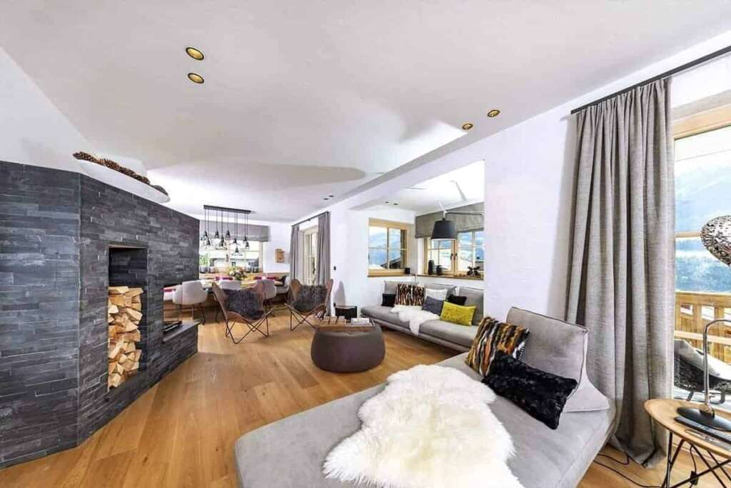 Offener Wohnraum mit grauer Steinkaminwand, Brennholz, hellem Sofa mit Fell und Essbereich