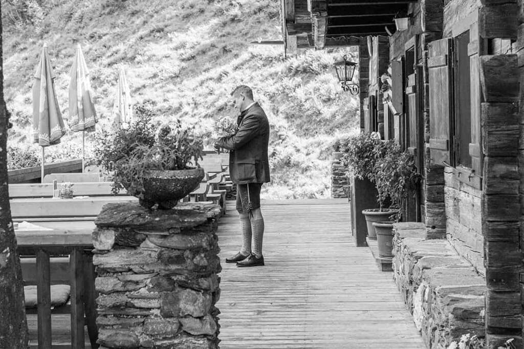 Schwarzweiß-Aufnahme eines Bräutigams in Tracht mit Blumenstrauß auf einer Holzterrasse vor altem Holzhaus
