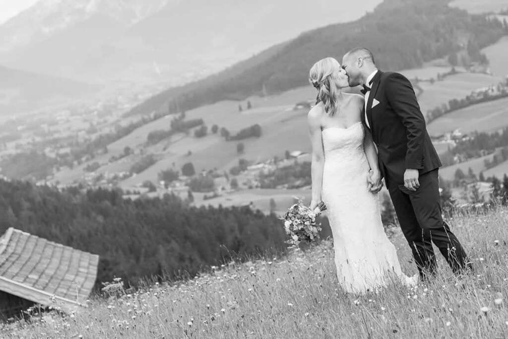 Schwarzweiß-Aufnahme eines sich küssenden Brautpaars auf einer Almwiese, im Hintergrund ein Tal mit Bergen
