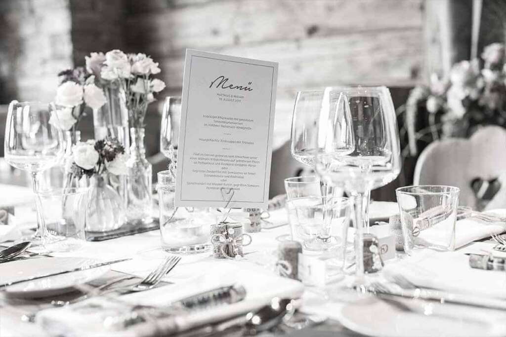 Schwarzweiß-Aufnahme einer gedeckten Hochzeitstafel mit Menükarte, Weingläsern und kleinen Blumenvasen