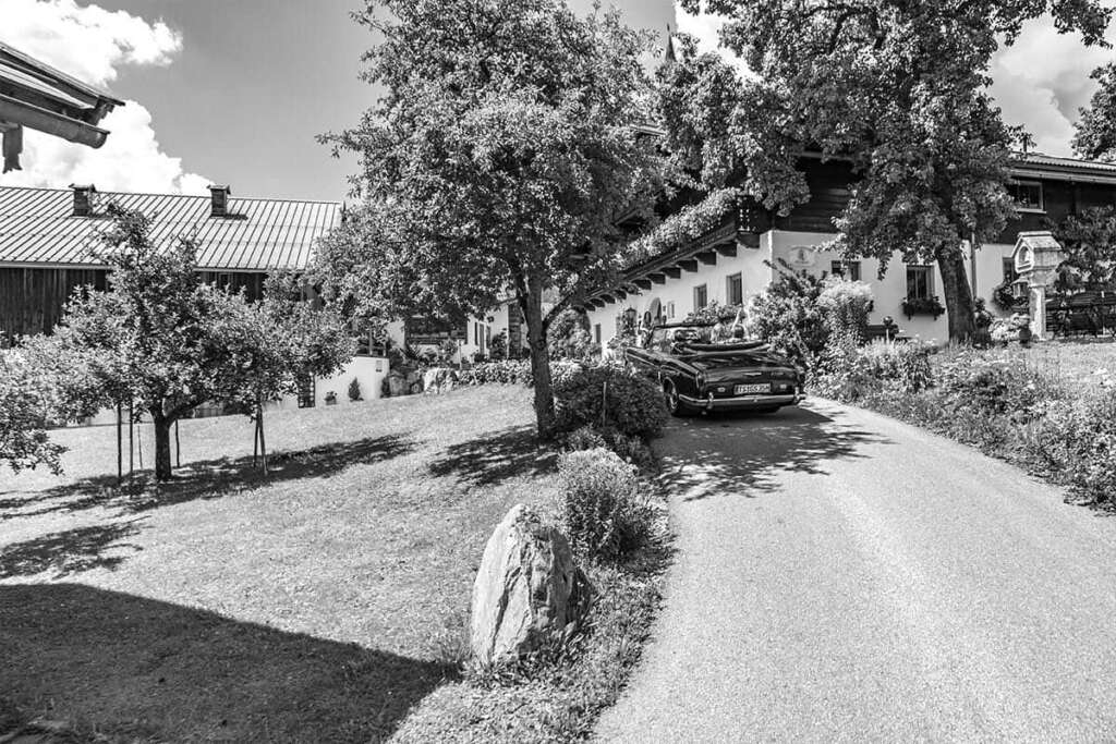 Schwarzweiß-Aufnahme eines Oldtimer-Cabrios auf der Zufahrt zu einem Bauernhof mit Bäumen und Garten