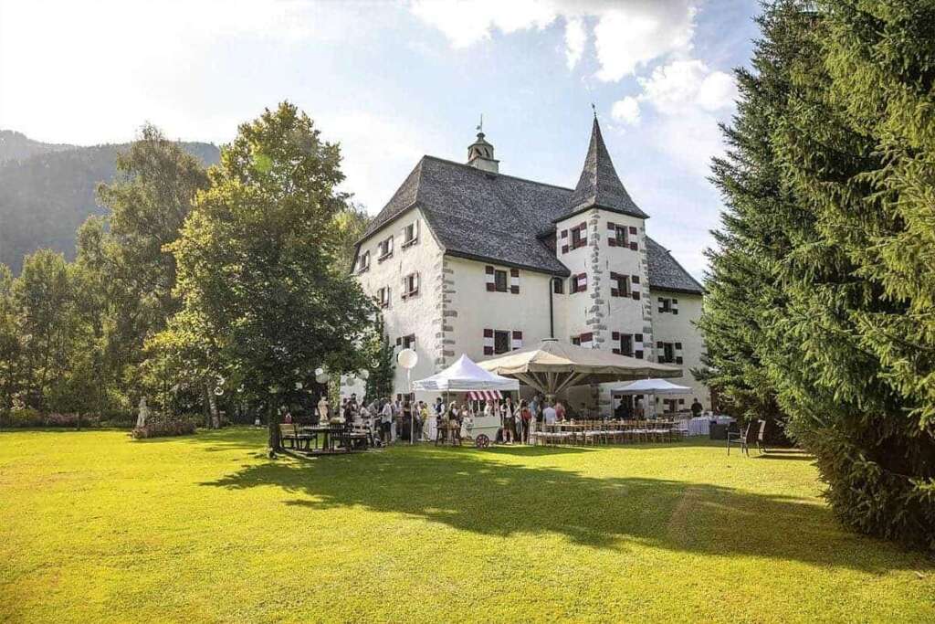 Sonniger Schlossgarten mit Rasen, Bäumen, weißen Zelten und Hochzeitsgästen vor einem Schloss mit Türmchen