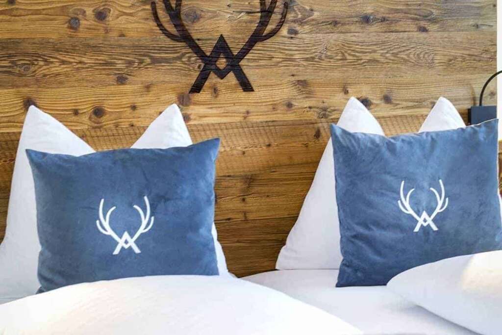 Blaue Kissen mit Geweihmotiv auf weißem Bett vor einer Altholzwand mit eingebranntem Geweihlogo