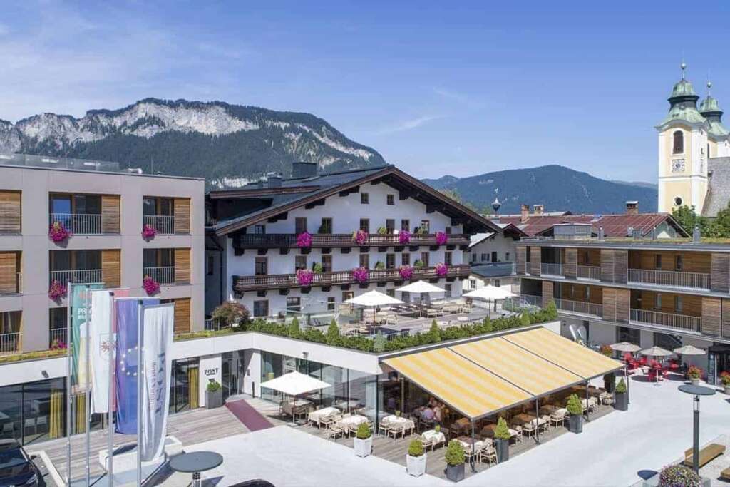 Hotel mit Gastgarten an einem Platz, dahinter Berge
