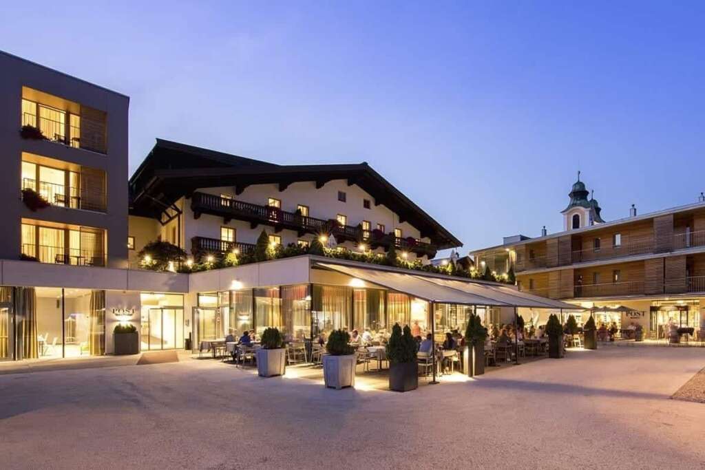 Hotel mit beleuchteter Terrasse und Gästen in der Abenddämmerung, dahinter ein Kirchturm