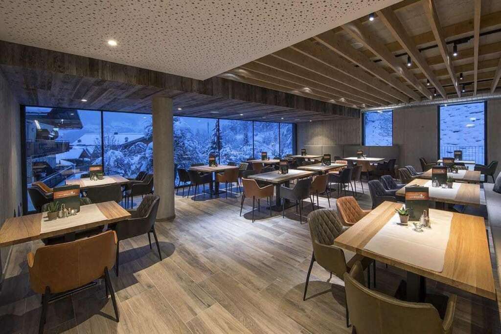 Modernes Restaurant mit Holztischen, Polsterstühlen und Fensterfront zu verschneiten Bäumen in der Dämmerung