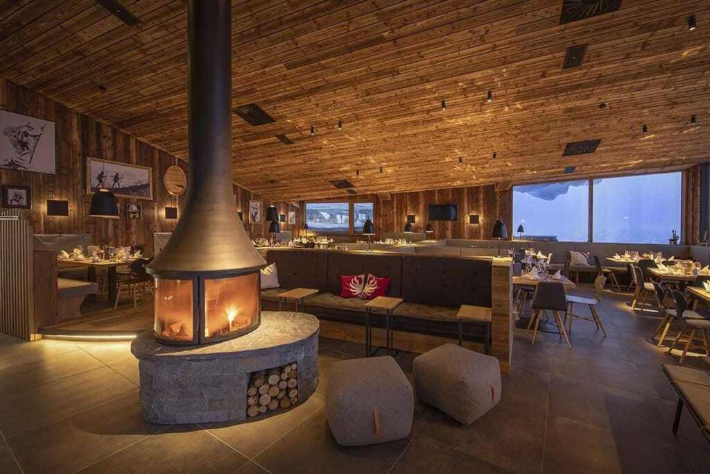 Restaurant mit Altholzdecke, frei stehendem Kamin mit Holzlager, gedeckten Tischen und Panoramafenstern