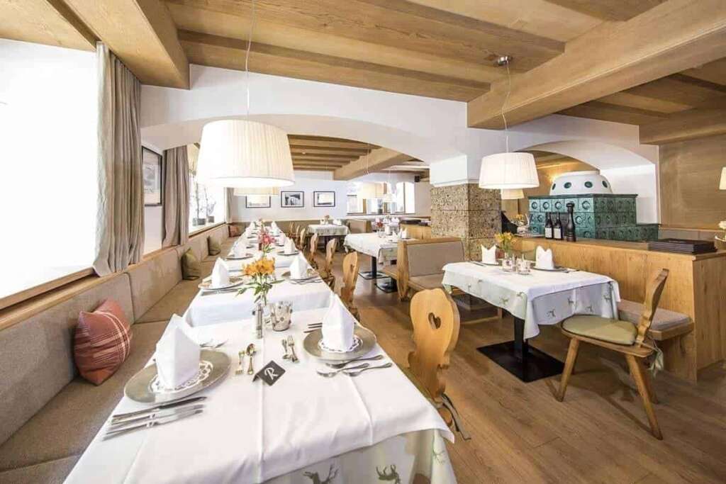 Helles Restaurant mit Holzbalkendecke, weiß gedeckter Tafel, Holzstühlen und grünem Kachelofen