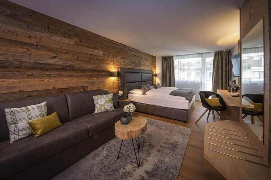 Hotelzimmer mit langer Altholzwand, dunklem Sofa, Baumscheibentisch, Doppelbett und Schreibtisch
