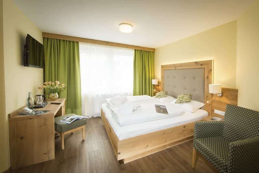 Hotelzimmer mit Holzbett, gepolstertem Kopfteil, grünen Vorhängen, gemustertem Sessel und Schreibtisch
