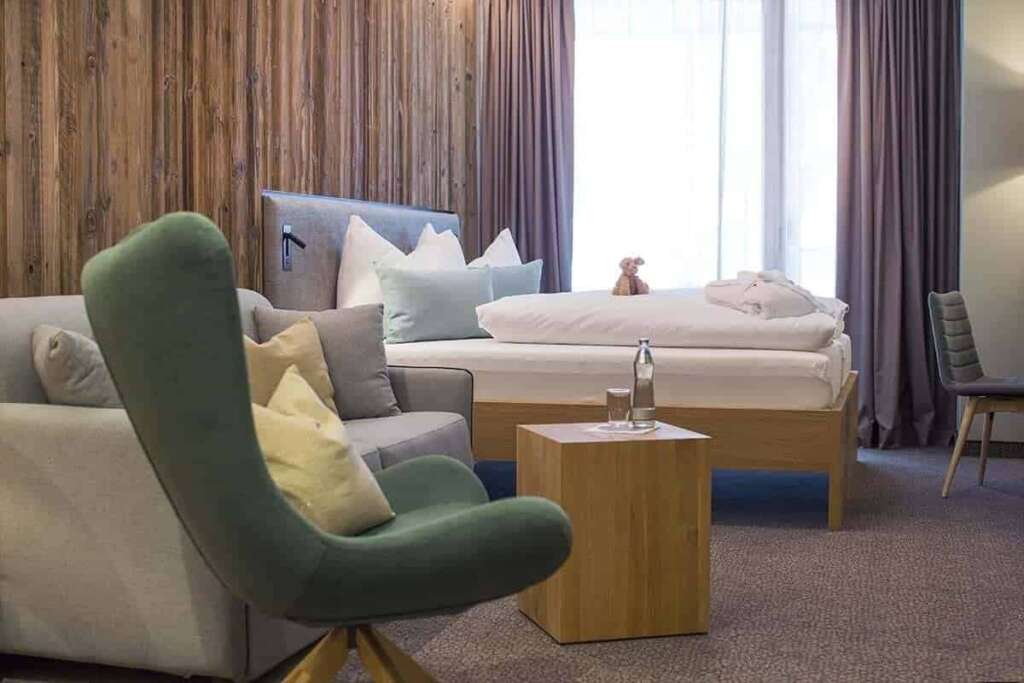 Hotelzimmer mit grünem Sessel, grauem Sofa, Holzwürfel, Bett und Altholzwand