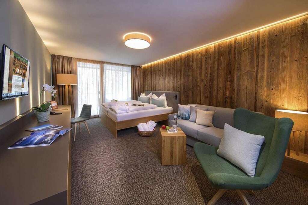 Hotelzimmer mit beleuchteter Altholzwand, grünem Sessel, Sofa, Doppelbett und Fernseher