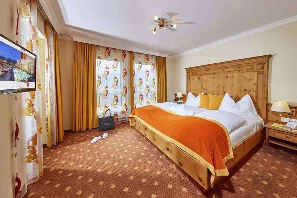 Hotelzimmer mit Holzbett, oranger Tagesdecke, gemusterten Vorhängen und Teppichboden