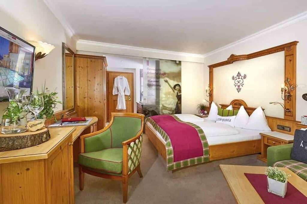 Hotelzimmer mit Holzbett, pinker Tagesdecke, grünem Sessel, Holzmöbeln und Bademantel an der Tür