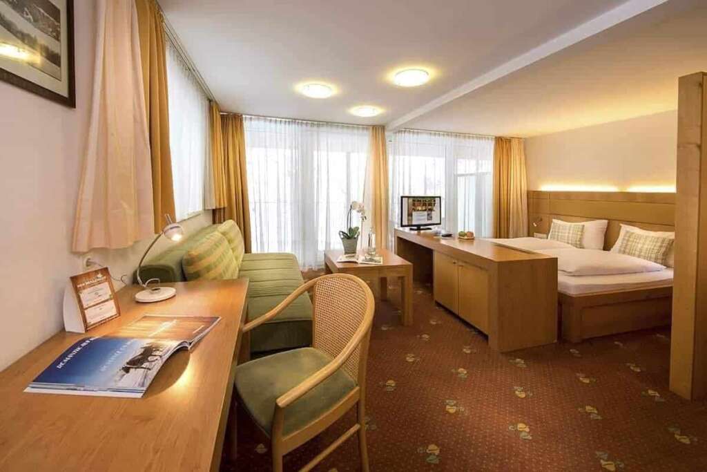 Hotelzimmer mit Holzschreibtisch, grünem Sofa, Doppelbett, rotem Teppichboden und hellen Vorhängen