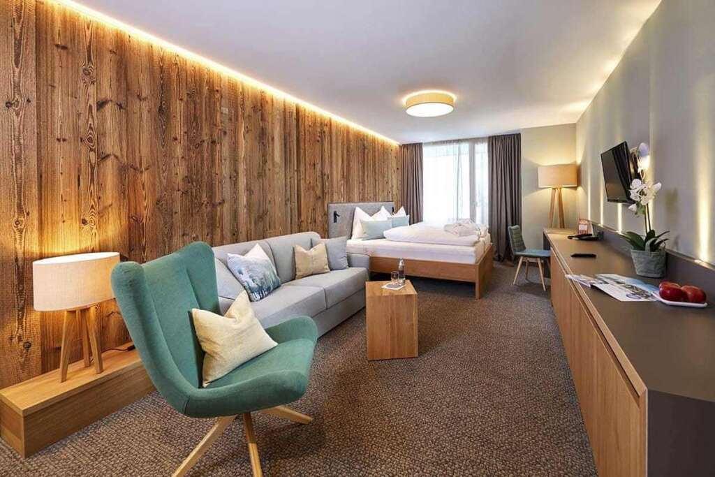 Helles Hotelzimmer mit türkisem Sessel, Sofa, Holzlampe, Bett und beleuchteter Altholzwand